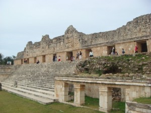 The image shows the ruins of an ancient stone structure, likely of Mayan origin, with a series of steps leading up to a long, rectangular building. The building has multiple doorways and intricate stone carvings on its facade. Several people are walking around and exploring the site. The sky is overcast, and the surrounding area is grassy.
