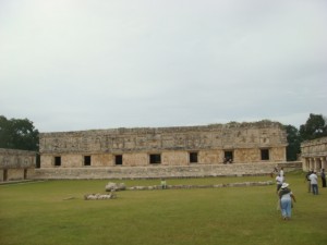 The image shows an ancient stone building with multiple doorways, likely part of a historical or archaeological site. The structure is rectangular and appears to be in a state of partial ruin. In the foreground, there is a grassy area with a few people walking and observing the site. The sky is overcast.