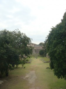 The image shows a pathway leading to an ancient stone structure, possibly a ruin, surrounded by lush greenery and trees. The sky above is cloudy, creating a somewhat overcast atmosphere. The pathway is grassy and appears to be well-trodden.