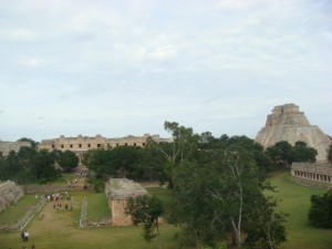The image shows an ancient archaeological site with several stone structures and ruins surrounded by greenery. In the background, there is a large pyramid-like structure. The sky is partly cloudy. There are a few people walking around the site, exploring the ruins.