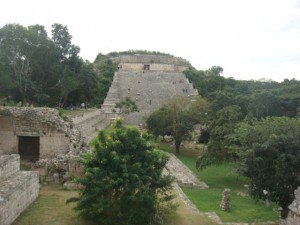 The image shows an ancient stone pyramid structure surrounded by lush greenery and trees. The pyramid has a series of steps leading up to the top, and there are ruins of other stone structures in the foreground. The scene appears to be part of an archaeological site, with a mix of natural and historical elements.