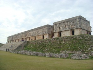 The image shows an ancient stone structure with intricate carvings, situated on a raised platform with a grassy area in the foreground. The building appears to be part of a historical or archaeological site, with steps leading up to the entrance. The sky above is cloudy.