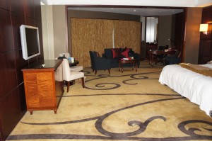 The image shows a spacious hotel room with a modern and elegant design. The room features a large bed with white linens on the right side, a flat-screen TV mounted on the wall to the left, and a wooden desk with a chair beneath the TV. The floor is covered with a beige carpet that has dark, swirling patterns. In the background, there is a sitting area with two dark-colored armchairs, a small table, and a desk with a chair. The room is well-lit with natural light coming from a window in the back.