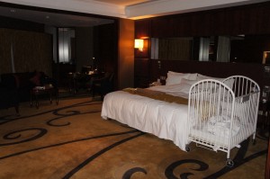 The image shows a spacious hotel room with a large bed covered in white linens and a brown throw blanket. Next to the bed is a white baby crib. The room has a warm, dim lighting with a lamp on the wall above the bed. The floor is carpeted with a design featuring swirls. In the background, there is a seating area with chairs and a table. The overall ambiance of the room is cozy and inviting.