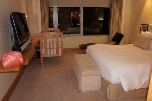 The image shows a hotel room with a large bed, a crib, a flat-screen TV mounted on the wall, and a cushioned bench at the foot of the bed. There is a window with curtains partially drawn, and a black chair with an ottoman near the window. A pink bag is placed on the desk below the TV. The room has a neutral color scheme with beige and white tones.