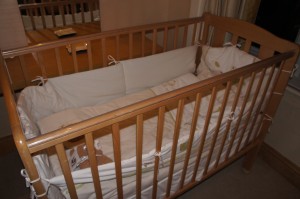 A wooden baby crib with white bedding and a mattress inside. The crib is placed in a room with a wooden floor and a window with curtains in the background. The crib has vertical slats on the sides and a solid headboard and footboard.
