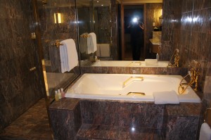 The image shows a luxurious bathroom with dark marble walls and flooring. There is a large white bathtub with gold fixtures, including a faucet and handles. The bathtub is built into a marble surround. Above the bathtub, there is a large mirror reflecting the room. On the left side, there are two white towels hanging on a towel rack. There are also some toiletries placed on the edge of the bathtub. The bathroom appears to be well-lit and spacious.