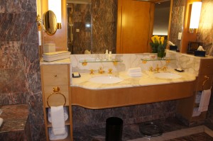 The image shows a luxurious bathroom with a double sink vanity. The vanity has a marble countertop and gold-colored fixtures. There are two mirrors above the sinks, each with a light fixture on the side. On the countertop, there are toiletries and a vase with yellow flowers. Below the sinks, there are shelves with neatly folded white towels. The bathroom has a polished stone wall and floor, and there is a black trash bin on the floor.