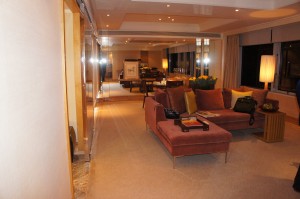 The image shows a spacious, well-lit living room with modern decor. There is a large, L-shaped sofa with a coffee table in front of it, which has some items on it. A floor lamp is placed next to the sofa, and there are large windows with curtains on the right side of the room. In the background, there is a dining area with a table and chairs. The room has a warm and inviting ambiance with soft lighting and neutral tones.