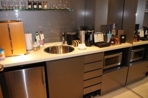 A modern kitchen countertop with various appliances and items. There is a sink in the center, surrounded by a coffee maker, a toaster, a microwave, and a few bottles and containers. Above the countertop, there is a glass shelf holding several glasses and bottles. The cabinets and drawers are sleek and metallic, giving the kitchen a contemporary look.