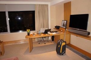 The image shows a modern hotel room or office space with a large window covered by curtains. There is a wooden desk with various items on it, including a laptop, a lamp, and some papers. A rolling suitcase is positioned next to the desk. On the wall, there is a large flat-screen TV mounted above a shelf. The room has a clean and organized appearance.