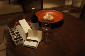 The image shows a round wooden table set with a bottle of red wine, two empty wine glasses, a box of chocolates, a corkscrew on a white napkin, and a wooden bowl containing bread rolls. The setting appears to be in a cozy, dimly lit room.