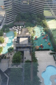 The image shows an aerial view of a recreational area situated between tall buildings. The area includes multiple swimming pools, a children's playground with colorful equipment, lounge chairs, and shaded seating areas. There is also greenery and landscaping throughout the space.