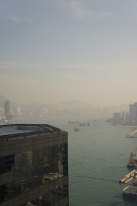 The image shows a hazy cityscape with a large body of water running through it. There are several boats on the water, and tall buildings on both sides of the waterway. The sky is overcast, contributing to the hazy atmosphere. In the foreground, there is a modern building with reflective windows. The background features more buildings and distant hills or mountains.