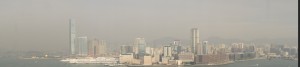 The image shows a panoramic view of a city skyline with numerous high-rise buildings. The city is situated along a waterfront, and the buildings are densely packed. The sky appears hazy, possibly due to fog or smog. The water in the foreground is calm, and there are mountains faintly visible in the background.
