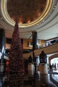 The image shows a large, elaborately decorated Christmas tree in the center of a spacious, luxurious hotel lobby. The tree is adorned with numerous lights and ornaments. The lobby features high ceilings with elegant lighting, tall black columns, and a grand staircase. There are a few people in the background, adding to the festive atmosphere.