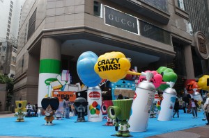 The image shows a festive outdoor display in front of a Gucci store. The display features large, colorful cartoon-like figures and objects, including a character holding a yellow balloon with the text "CRAZY X'MAS!" written on it. The ground is covered with a bright blue material, and there are various other decorations and figures around, creating a lively and playful atmosphere. The surrounding buildings and cityscape are visible in the background.