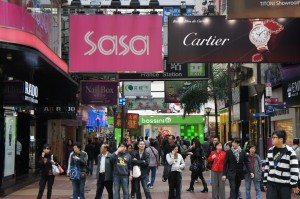 The image shows a busy shopping street with numerous people walking and shopping. There are various store signs visible, including prominent ones for "Sasa" and "Cartier." The street is lined with shops and advertisements, and the atmosphere appears lively and bustling.