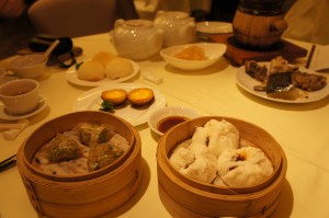 The image shows a table set with various dim sum dishes. There are bamboo steamers containing dumplings and buns, a plate with egg tarts, a bowl with a yellowish dessert, a teapot, and cups of tea. The table is covered with a light-colored tablecloth, and the setting appears to be in a restaurant.