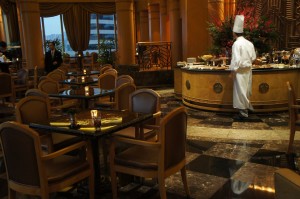 The image shows an elegant restaurant interior with a chef in a white uniform and hat standing behind a circular counter. The restaurant has several tables and chairs arranged neatly, with some tables set with candles. The floor is made of polished marble tiles, and the background features large windows with draped curtains, allowing natural light to enter. There are also decorative elements and plants enhancing the ambiance.