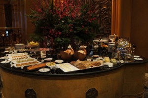 The image shows an elegant buffet table set up with a variety of food items. The table includes an assortment of breads, pastries, cheeses, and other appetizers. There are also several small dishes and plates arranged neatly around the table. In the background, there is a large floral arrangement with red and green foliage, adding a decorative touch to the setting. The overall ambiance suggests a luxurious and well-organized dining experience.