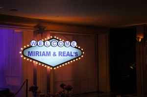 The image shows a lit-up sign in a dark setting. The sign is designed in a retro style with a diamond shape and is surrounded by small lights. It reads "WELCOME MIRIAM & REAL'S" in capital letters. The background appears to be an indoor space with curtains and some plants visible at the bottom of the image.