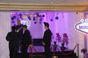 A group of people dressed in formal attire are standing near a stage decorated with purple lighting and floral arrangements. There is a red rope barrier in front of the stage, and a sign with the word "MIR" is partially visible on the right side of the image. The setting appears to be an event or a formal gathering.