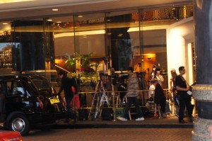 A group of people is gathered outside a building with large glass windows. There is a black vehicle parked nearby, and various equipment, including tripods and lights, is visible. The scene appears to be busy, possibly indicating a film or photo shoot taking place. The building's interior is well-lit, and some greenery can be seen inside.