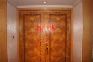 A set of ornate wooden double doors with intricate patterns. The doors have two red Chinese characters, one on each door, which are often associated with good luck or happiness. The surrounding walls are light-colored with a textured pattern, and there is a small light fixture on the ceiling above the doors.