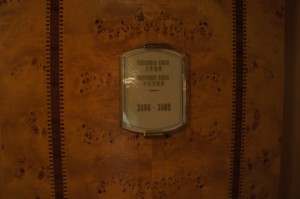 The image shows a wooden wall with a plaque mounted on it. The plaque has some text on it, but the text is not clearly readable due to the lighting and the angle of the photograph. The wood has a rich, warm tone with a natural grain pattern. The plaque appears to be metallic or reflective, and it is framed in a way that makes it stand out against the wood.