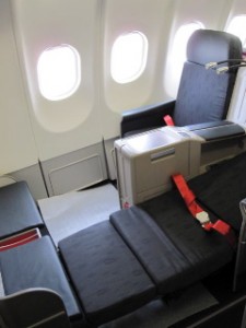 The image shows a business class seat in an airplane. The seat is fully reclined into a flatbed position. It has a black cushion and red seatbelt. The seat is located next to the airplane windows, and there is a small console with storage space and a tray table. The overall setting appears to be spacious and designed for comfort during long flights.