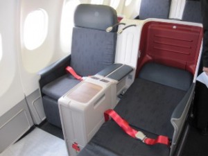 The image shows a business class seat on an airplane. The seat is spacious and includes a footrest, a reclining backrest, and a red seatbelt. There is a partition between the seats, and the area appears to be well-lit with natural light coming from the windows on the left side. The seat is upholstered in dark fabric, and there is a small storage compartment and a tray table in the center console.