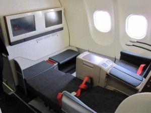 The image shows the interior of a first-class or business-class airplane cabin. The cabin features two spacious seats that can be converted into flat beds, each with a red seatbelt. There are two windows on the side, and the seats are equipped with personal entertainment screens. The area appears to be designed for comfort and privacy, with ample legroom and a modern, sleek design.