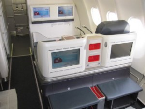 The image shows the interior of an airplane's business or first-class section. There are two large, comfortable seats with individual entertainment screens in front of them. The seats are separated by a partition that also houses additional screens and storage compartments. The cabin appears spacious and well-lit, with windows on the right side. The overall design is modern and geared towards providing a comfortable and private experience for passengers.