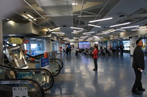 The image shows the interior of an airport terminal. There are people walking and sitting in the waiting area. An escalator is visible on the left side of the image, and a large digital flight information board can be seen in the background. The ceiling has a modern design with various light fixtures. The overall atmosphere appears to be calm and organized.