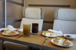 The image shows a dining setup on a table with two cushioned chairs in the background. The table has two plates of food, each with a small sandwich, a piece of cake, and a pastry. There are also two drinks, one light-colored and one dark-colored, placed in the center of the table. White napkins and cutlery are neatly arranged beside the plates. The setting appears to be in a comfortable, possibly luxurious environment.