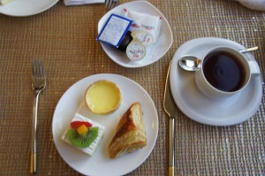 A breakfast setting is shown in the image. There is a white plate with a small tart, a piece of cake topped with fruit, and a pastry. Next to the plate is a cup of black coffee on a saucer with a spoon. Above the plate, there is a small dish containing packets of butter and jam. A fork is placed to the left of the plate, and a knife is to the right of the coffee cup. The table is covered with a woven placemat.