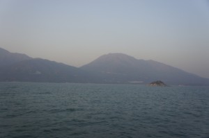 The image shows a calm body of water with a small rocky island in the middle distance. In the background, there are large, hazy mountains under a clear sky. The overall atmosphere is serene and peaceful.