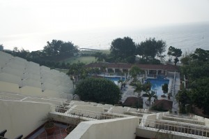 The image shows a view from a higher vantage point overlooking a resort area. In the foreground, there are multiple levels of white buildings with staircases and railings. Below, there is a large swimming pool surrounded by lounge chairs and umbrellas. Beyond the pool area, there are trees and greenery, and in the background, the ocean is visible under a hazy sky.