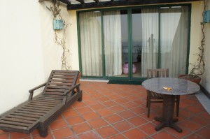 The image shows a small outdoor patio area with a tiled floor. There is a wooden lounge chair on the left side and a round table with two chairs on the right side. The table has a small object on it, possibly a piece of fruit or a snack. The patio is adjacent to a building with large sliding glass doors covered by light-colored curtains. There are some decorative elements, including a lantern and climbing plants on the wall.