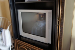 The image shows an older model CRT television placed inside a black entertainment center. To the left of the television, there is a white baby crib with some white fabric draped over its side. The entertainment center has a decorative design on its lower section. The reflection on the television screen shows a bed and a lamp in the background.