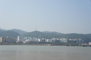 The image shows a coastal cityscape with a row of buildings along the shoreline. In the background, there are green hills with a few structures, including a tall antenna or tower, on top of them. The sky is clear and blue, and the water in the foreground is calm.