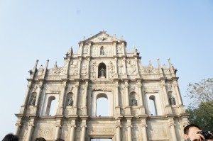The image shows the facade of the Ruins of St. Paul's, a historic landmark in Macau. The structure is a stone facade with intricate carvings and statues, standing tall against a clear blue sky. There are some people visible at the bottom of the image, partially cut off. The facade features multiple arched windows and detailed architectural elements.
