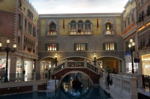 The image shows an indoor scene designed to resemble a Venetian canal. There is a small arched bridge over a canal with a gondola passing underneath. The surrounding architecture features buildings with balconies and arched windows, reminiscent of Venetian style. The area is well-lit with street lamps and there are shops visible on either side of the canal. The ceiling is painted to look like the sky, giving the impression of an outdoor setting.