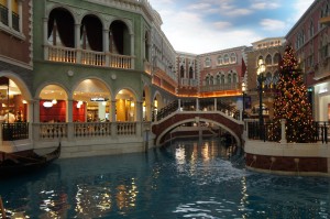 The image shows an indoor scene designed to resemble a Venetian canal. There is a waterway with a gondola on the left side, arched bridges, and buildings with arched windows and balconies. The buildings are decorated with festive lights, and there is a large Christmas tree on the right side of the image. The ceiling is painted to look like a blue sky with clouds.