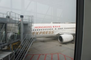 The image shows a Hong Kong Express airplane parked at an airport gate. The aircraft is connected to the terminal via a jet bridge. The plane's fuselage is white with "HONG KONG" written in red and "EXPRESS" written in gold. The scene is viewed through a window from inside the terminal.