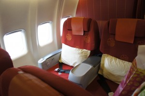 The image shows two adjacent airplane seats by the window. The seats are upholstered in red fabric with a pattern and have headrests covered with red cloth. There are white pillows placed on each seat. The window shades are open, allowing light to enter the cabin. The armrest between the seats has a built-in tray and a seatbelt is visible on one of the seats.