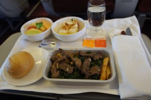 The image shows an airplane meal served on a tray. The meal includes a main dish of beef strips with vegetables and possibly potatoes, a side salad with slices of melon and a leaf of mint, a dessert that appears to be a piece of cake with a topping, a bread roll on a small plate, a glass of water, and a set of utensils wrapped in a white napkin. There are also two small packets, likely containing condiments or seasonings, placed next to the main dish.