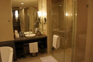 The image shows a modern bathroom with a glass-enclosed shower on the right side. There is a white towel hanging on the shower door. To the left of the shower is a vanity area with a round mirror, a sink, and various toiletries on the countertop. Below the countertop, there are two white towels hanging. The bathroom has a clean and organized appearance with neutral-colored walls and flooring. There is also a bathtub partially visible on the left side of the image.