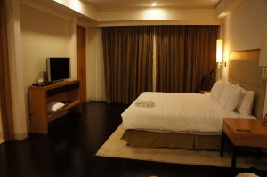 The image shows a modern hotel room with a large bed covered in white linens and several pillows. There is a wooden nightstand with a lamp on one side of the bed. A flat-screen TV is mounted on a wooden stand against the wall opposite the bed. The room has dark wooden flooring and a light-colored area rug under the bed. The far wall features large windows covered with dark curtains. The lighting in the room is warm and ambient.